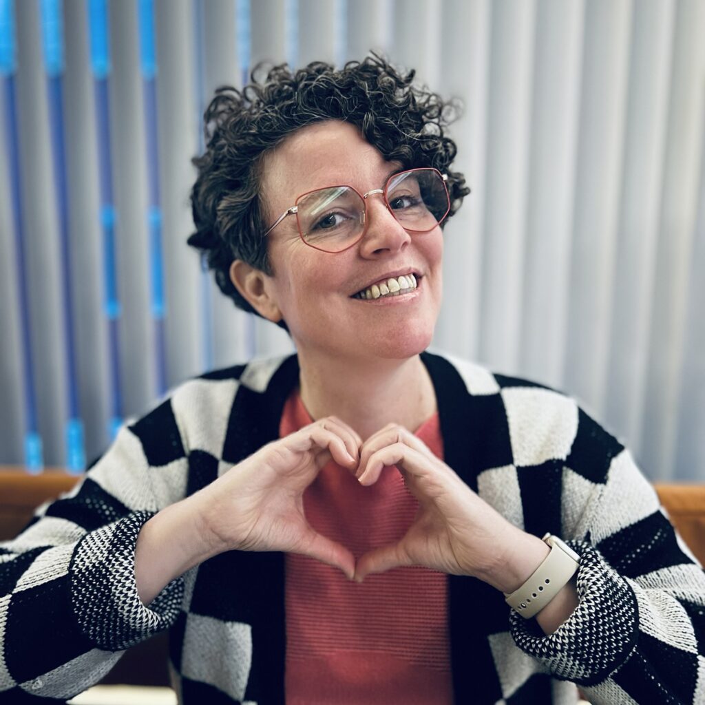 Jill Murray smiles and makes a heart shape with her hands. She has short hair and glasses, and wears a black and white check cardigan over a bright pink top.